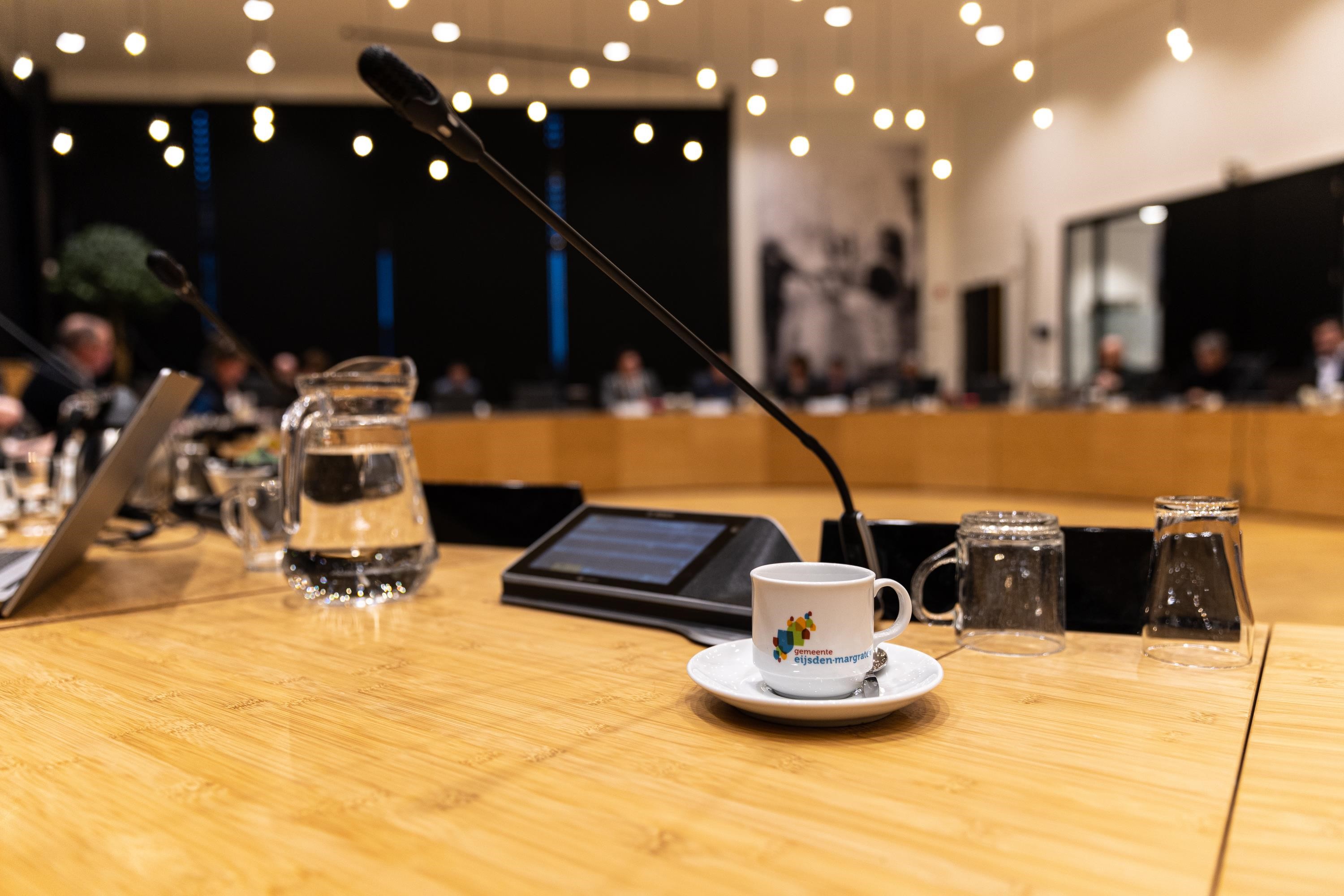 Raadzaal in ovale opstelling houten tafels met in beeld een microfoon, kan water en glas en een koffiekopje met logo gemeente Eijsden-Margraten
